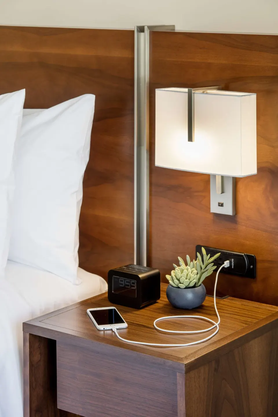Bedside table with lamp, clock, phone, and headboard in a suite at Hilton Garden Inn Waikiki Beach.