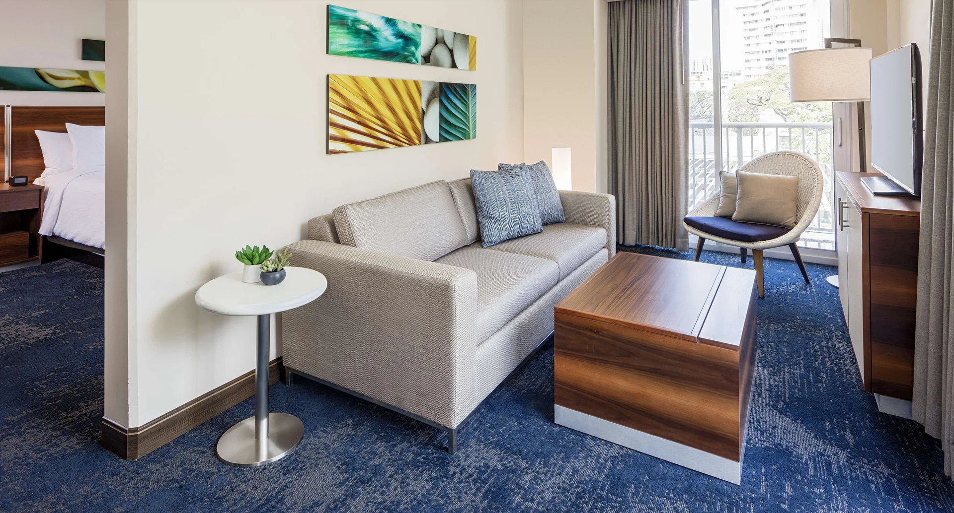 Living room with sofa, chair, coffee table, balcony door, and bedroom beyond at Hilton Garden Inn Waikiki Beach.