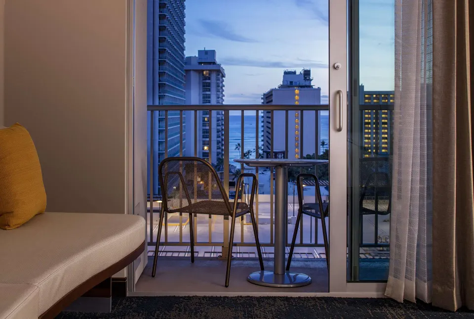 Standard Room balcony seating with city buildings and ocean beyond at Hilton Garden Inn Waikiki Beach.