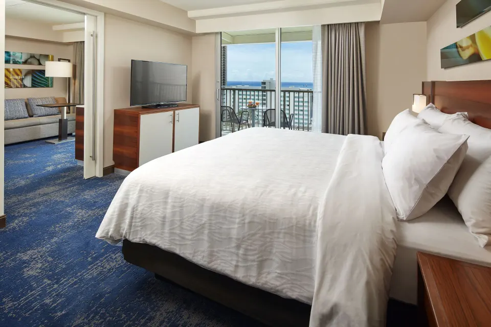 Accessible suite king bed, sitting area, balcony, and ocean glimpse at Hilton Garden Inn Waikiki Beach.