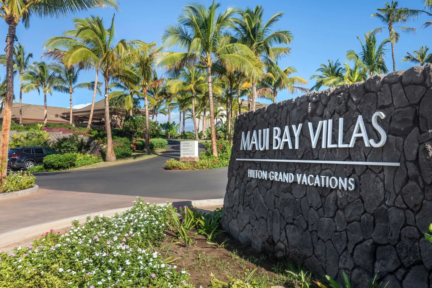 Entrance sign at Hilton Grand Vacations Club Maui Bay Villas set among palms, flowers, and a driveway.