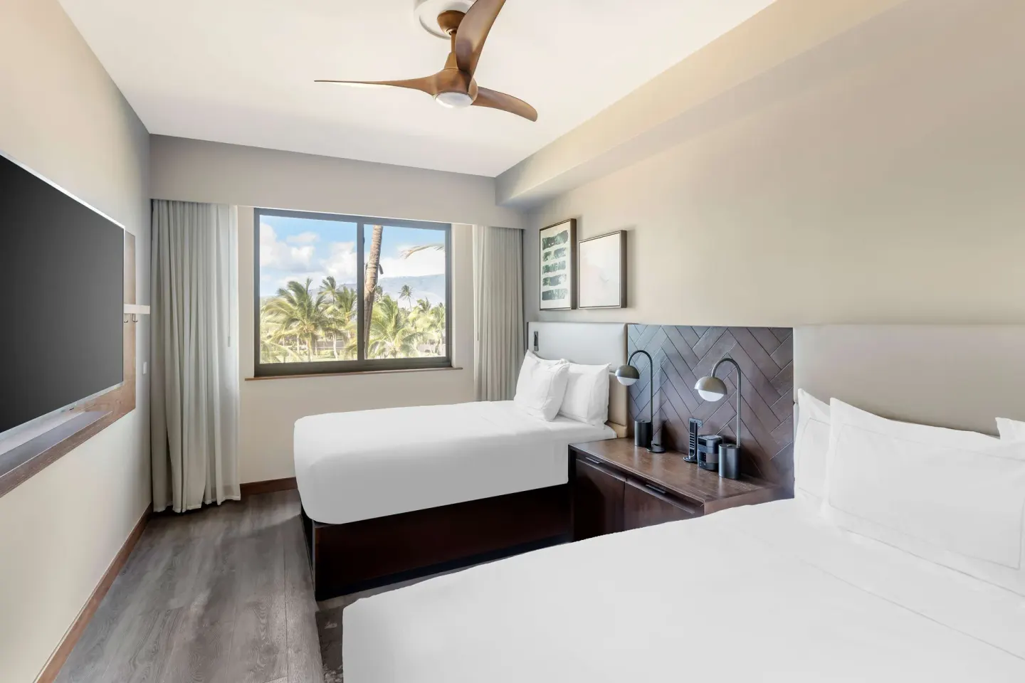 Two white beds, wall-mounted TV and palm-view window in a suite at Hilton Grand Vacations Club Maui Bay Villas.