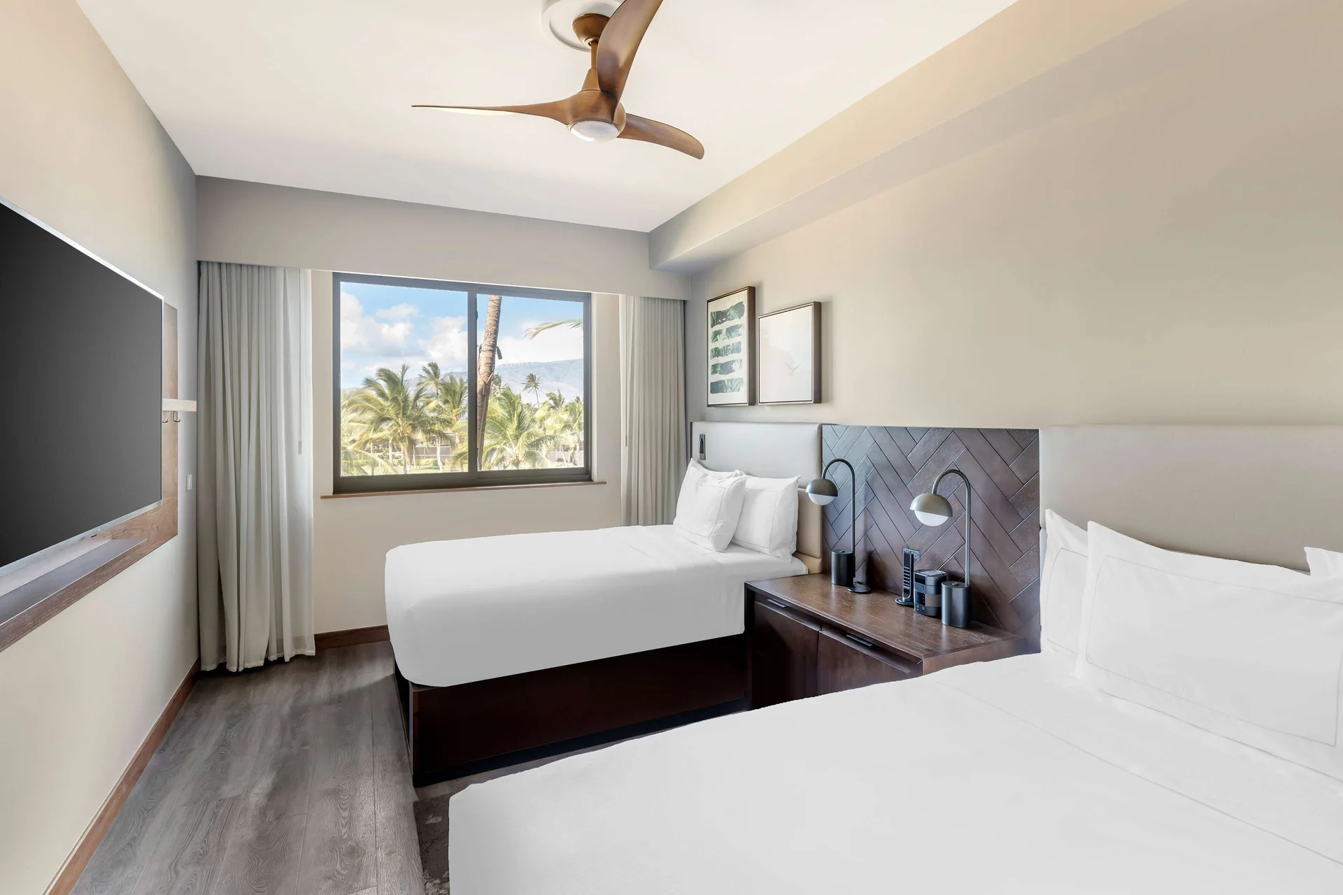 Two white beds, wall-mounted TV and palm-view window in a suite at Hilton Grand Vacations Club Maui Bay Villas.
