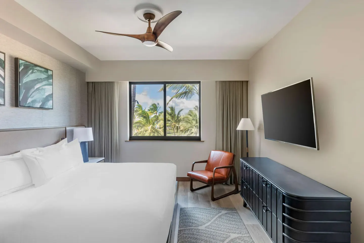 King bedroom with wall TV, leather chair, and palm-framed window at Hilton Grand Vacations Club Maui Bay Villas.