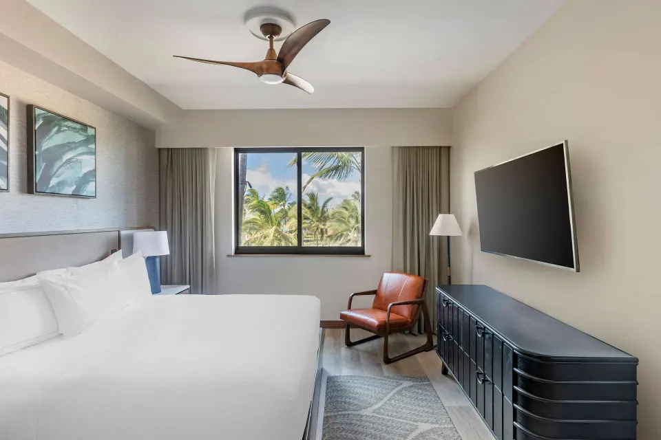 King bedroom with wall TV, leather chair, and palm-framed window at Hilton Grand Vacations Club Maui Bay Villas.
