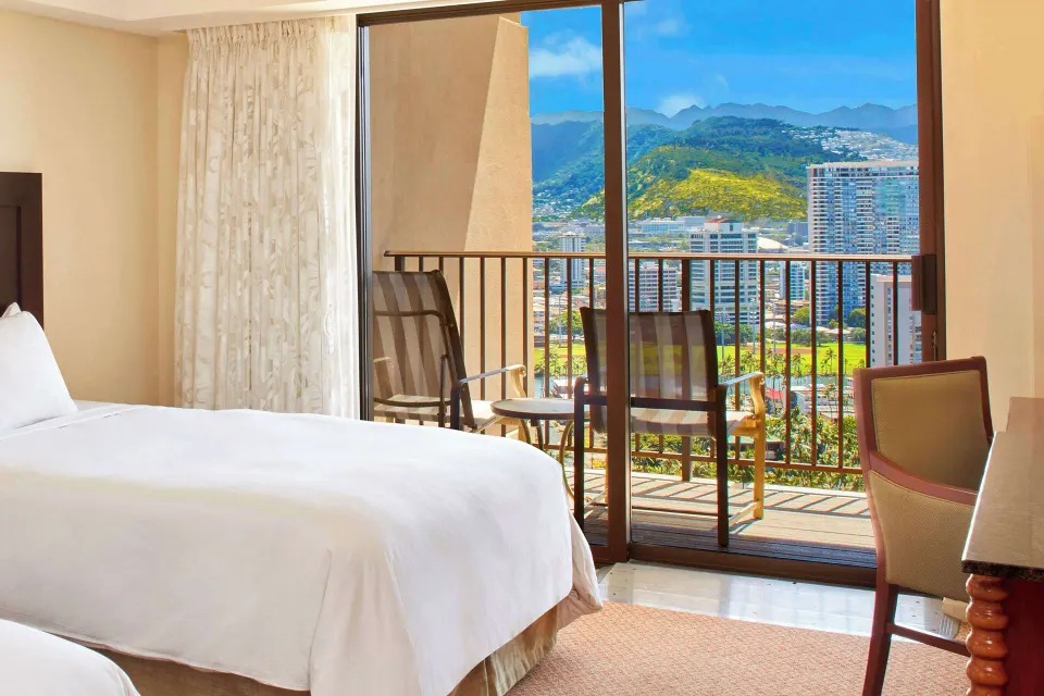 Resort view room with white bed and balcony overlooking mountains at Hilton Hawaiian Village Waikiki Beach Resort.