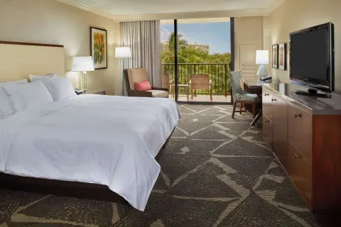 Resort view room with king bed, TV, desk, and palm-view balcony at Hilton Hawaiian Village Waikiki Beach Resort.