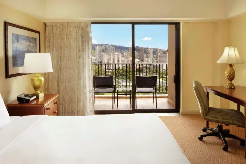 Resort view room with bed, desk, lamps, and city balcony at Hilton Hawaiian Village Waikiki Beach Resort.