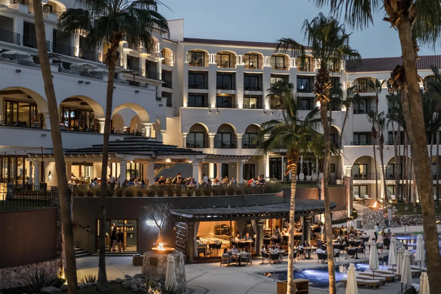 Evening view of illuminated resort buildings, terraces, palms and poolside dining at Hilton Los Cabos.