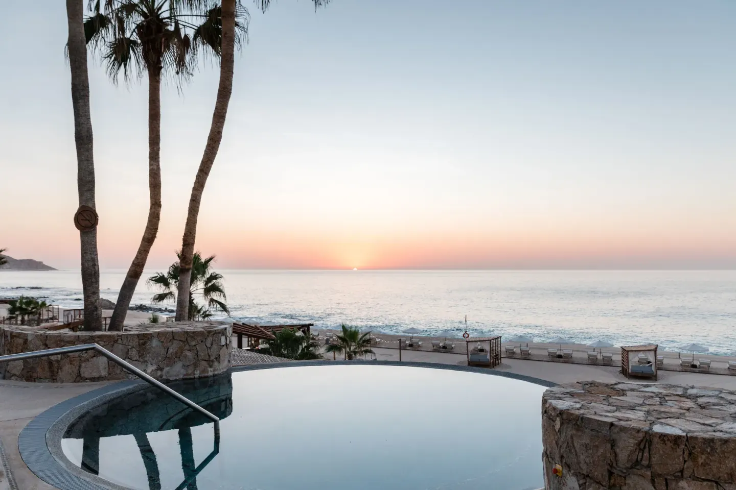 Sunset over the ocean beyond a round pool and palms at Hilton Los Cabos.