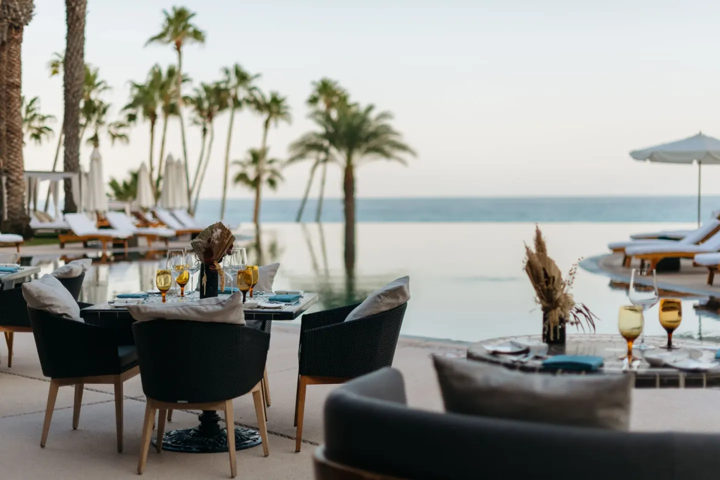 Poolside dining tables set beside palm trees and the ocean at Hilton Los Cabos.