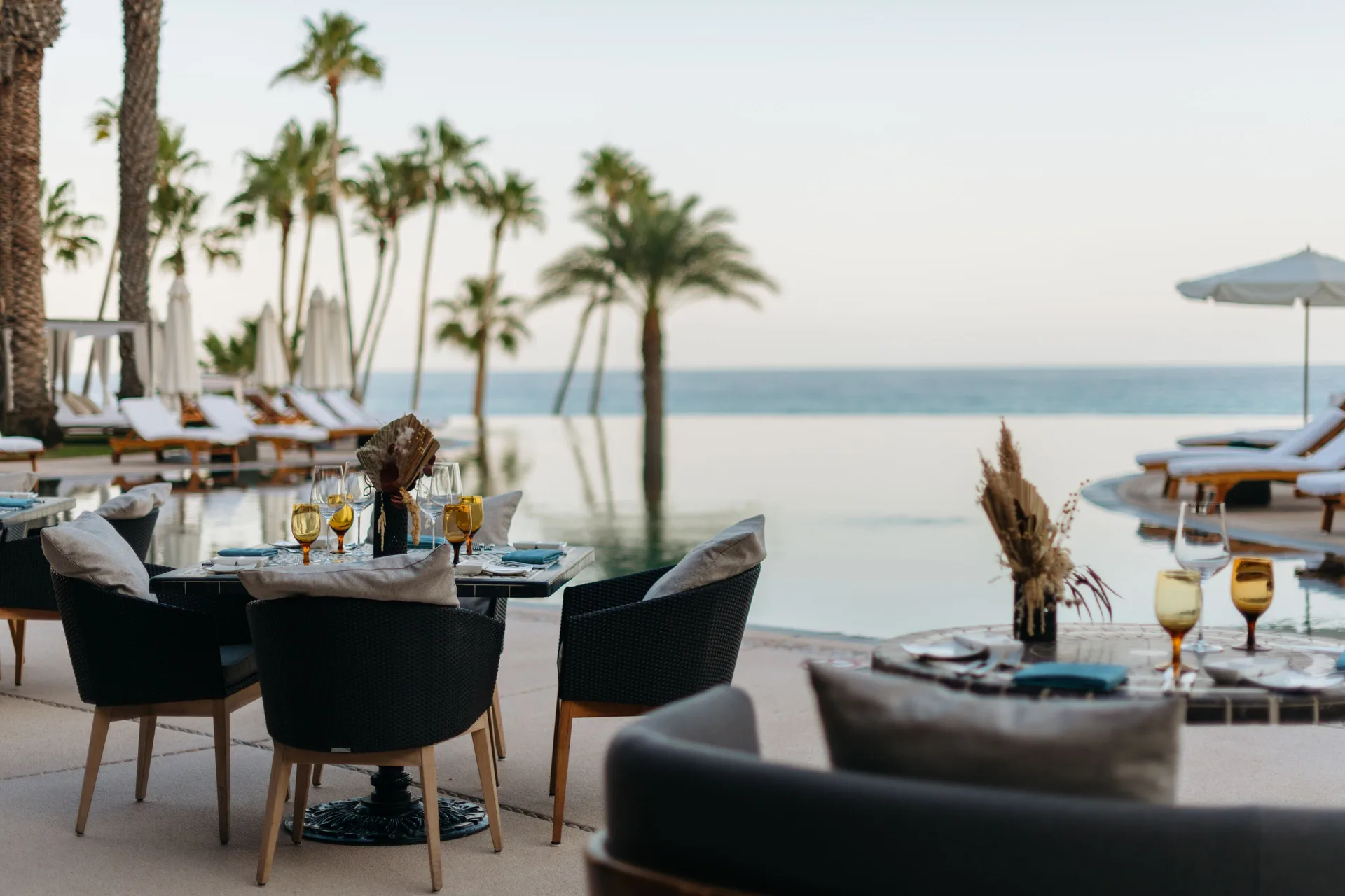 Poolside dining tables set beside palm trees and the ocean at Hilton Los Cabos.