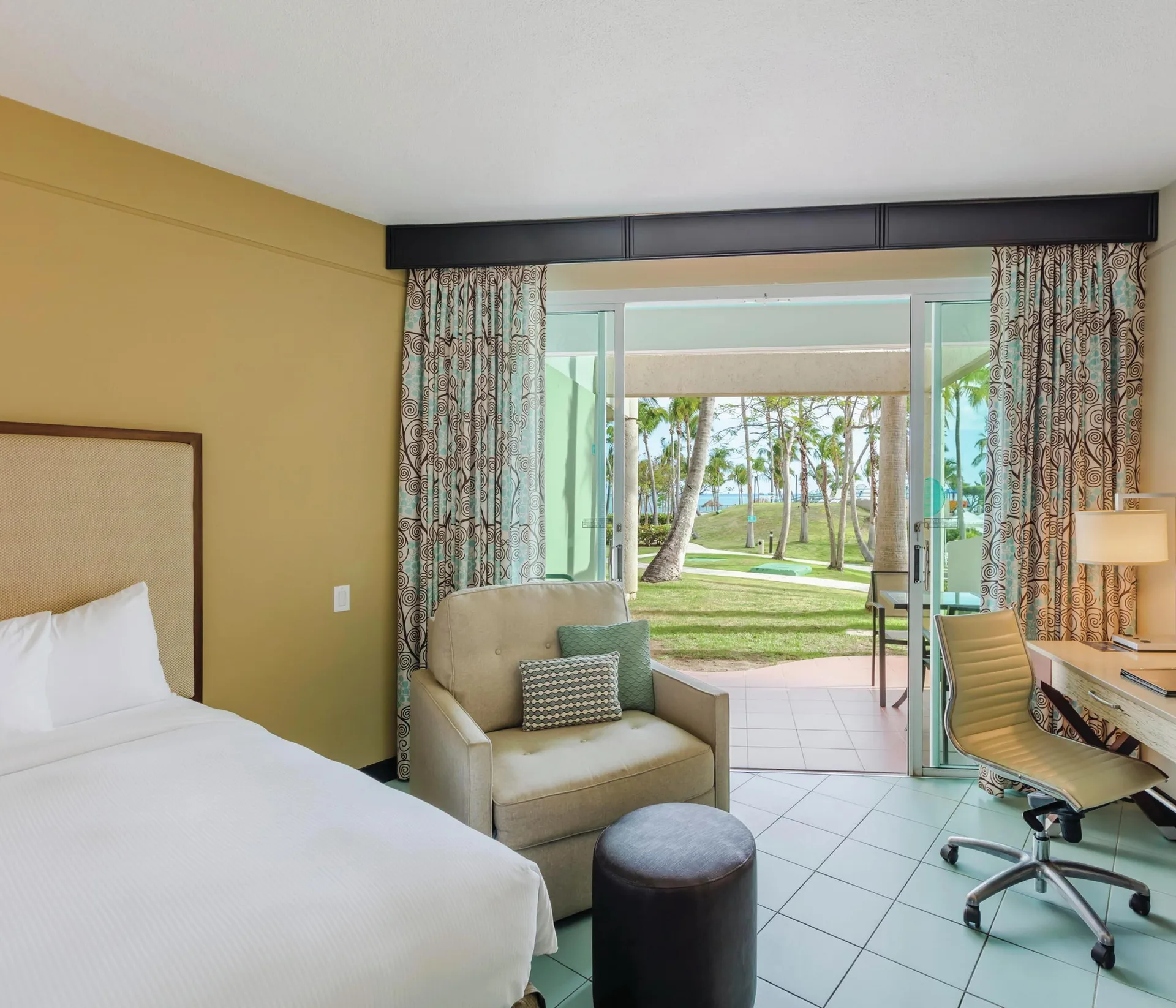 Oceanfront patio room at Hilton Ponce Golf & Casino Resort with partial bed, chair, desk, and lawn view.