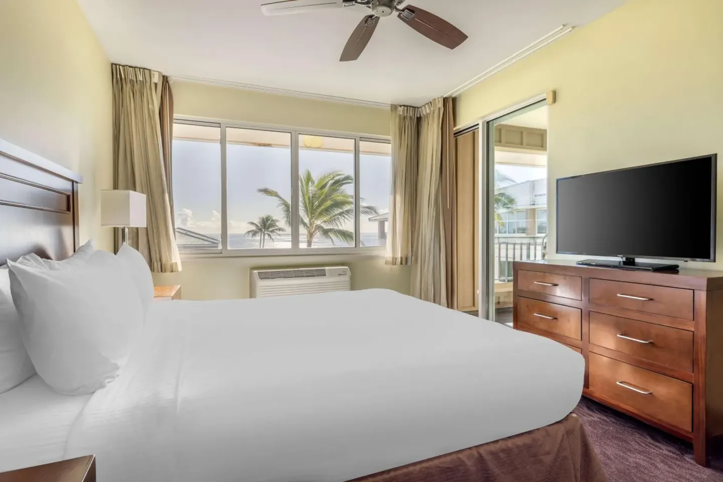 Guest room with king bed, dresser, TV, and balcony at Hilton Vacation Club The Point At Poipu Kauai.