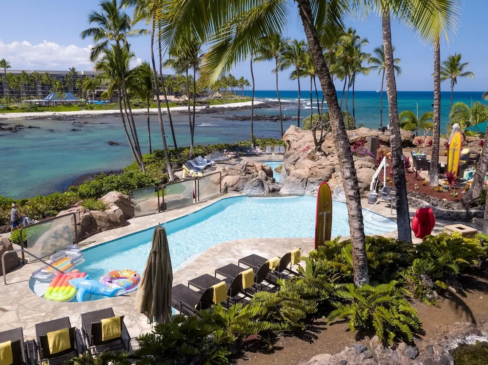 Oceanfront pool with water slide, floats, palms, and lagoon view at Hilton Waikoloa Village.