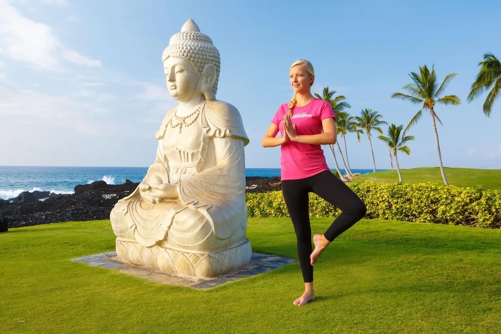 Woman practices yoga beside a seated statue and ocean lawn at Hilton Waikoloa Village.