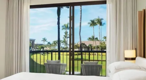 Guest room bed beside a balcony with palm and lawn views at Hilton Waikoloa Village.