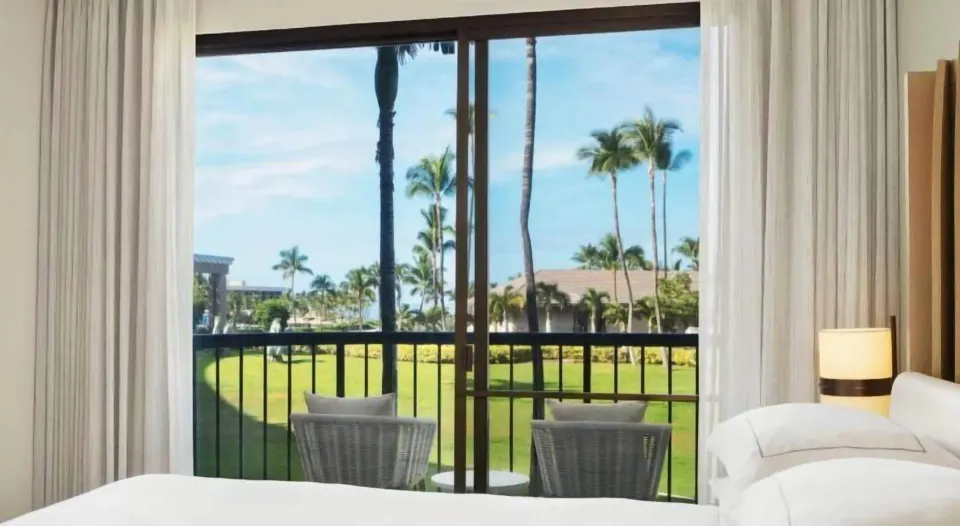 Guest room bed beside a balcony with palm and lawn views at Hilton Waikoloa Village.