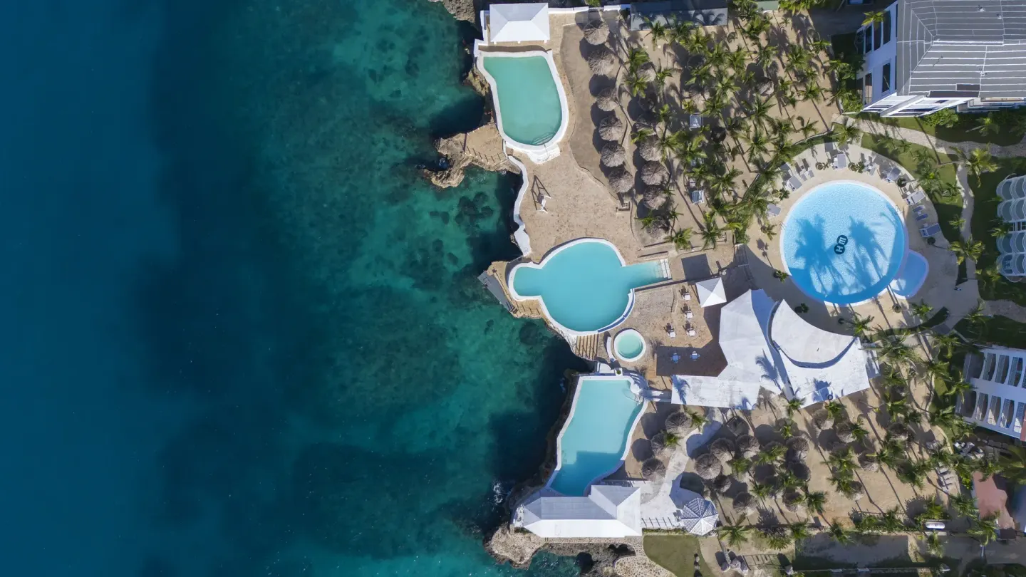 Top-down aerial view of pools, palms, and clear sea at HM Alma de Bayahibe.