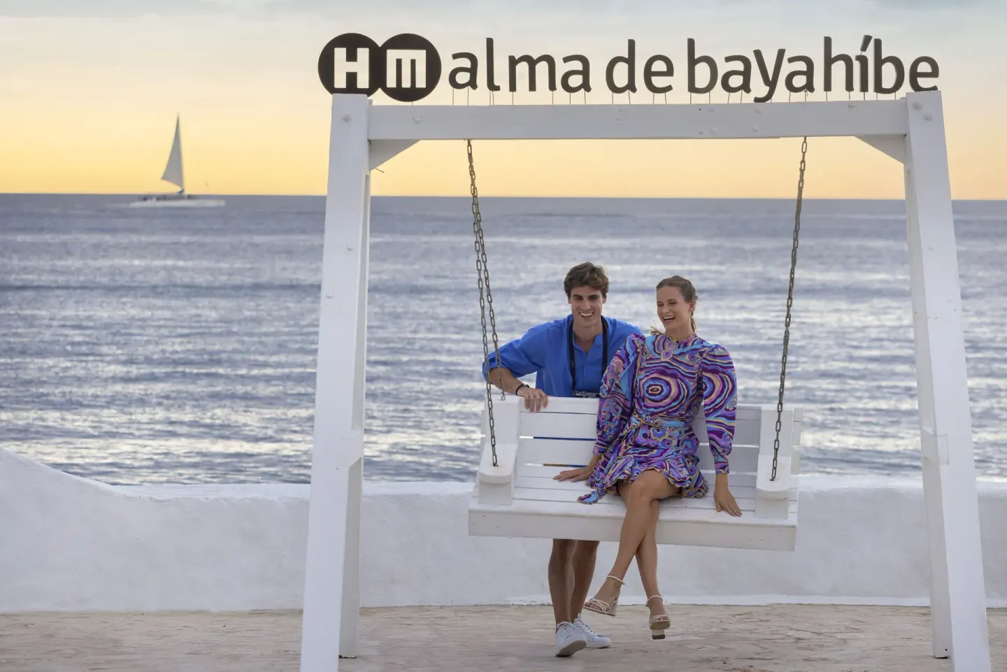 Couple seated on a white oceanfront swing with HM Alma de Bayahibe sign above.