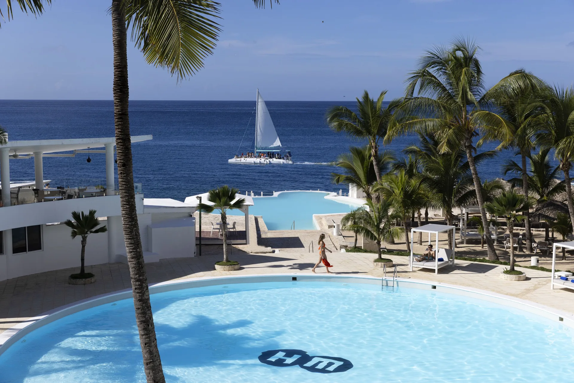 Palm-framed pools and a sailing catamaran offshore at HM Alma de Bayahibe.