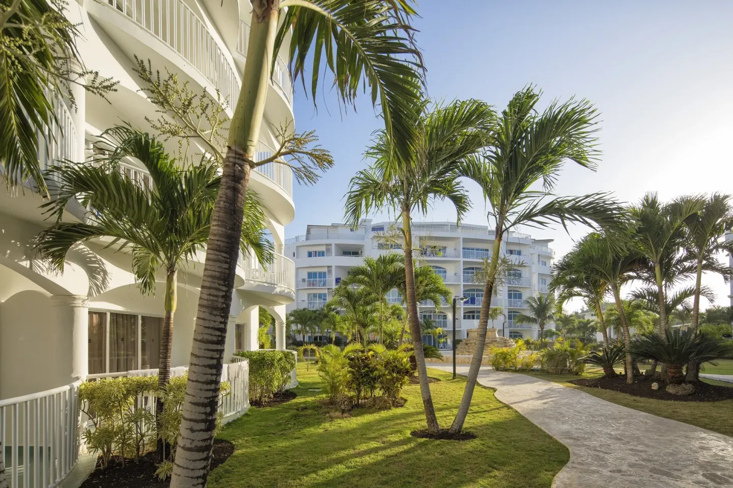 Palm-lined garden walkway between white guest buildings at HM Alma de Bayahibe.