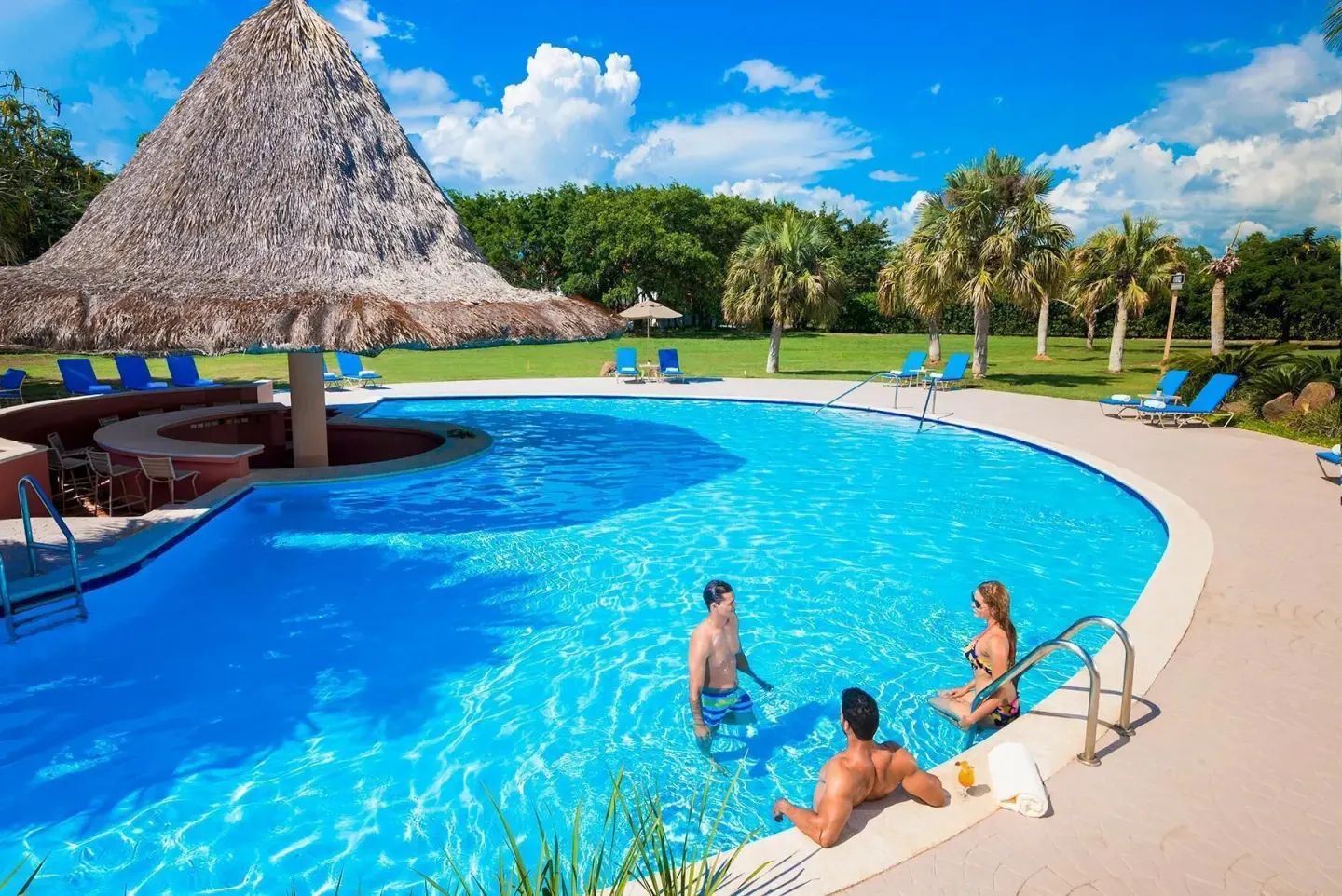 Guests relax in a bright pool beside a thatched bar at Hodelpa Garden Suites Golf & Convention Center.
