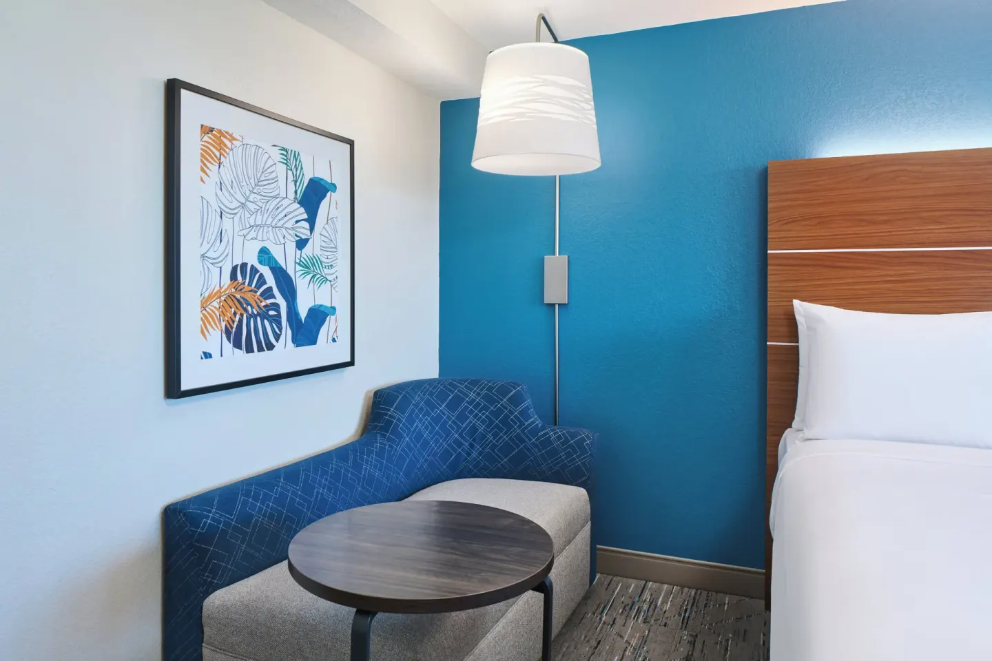 Standard Room king bed corner with blue chair, round table, and wall art at Holiday Inn Express Waikiki.