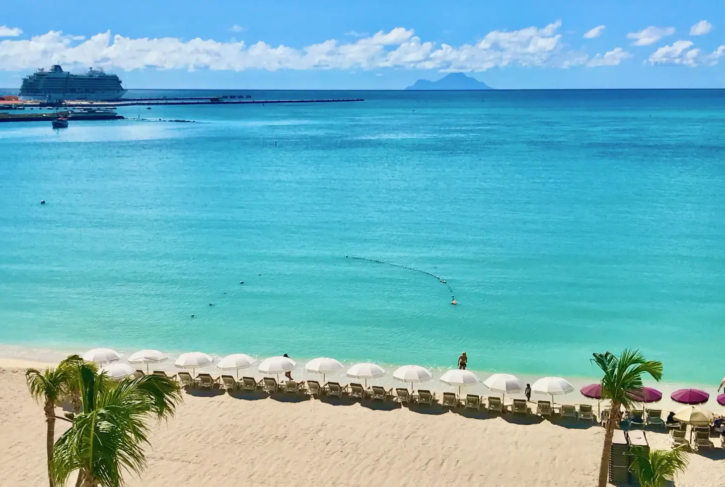 Beach view at Holland House Beach Hotel with white umbrellas, turquoise water, and a cruise ship.