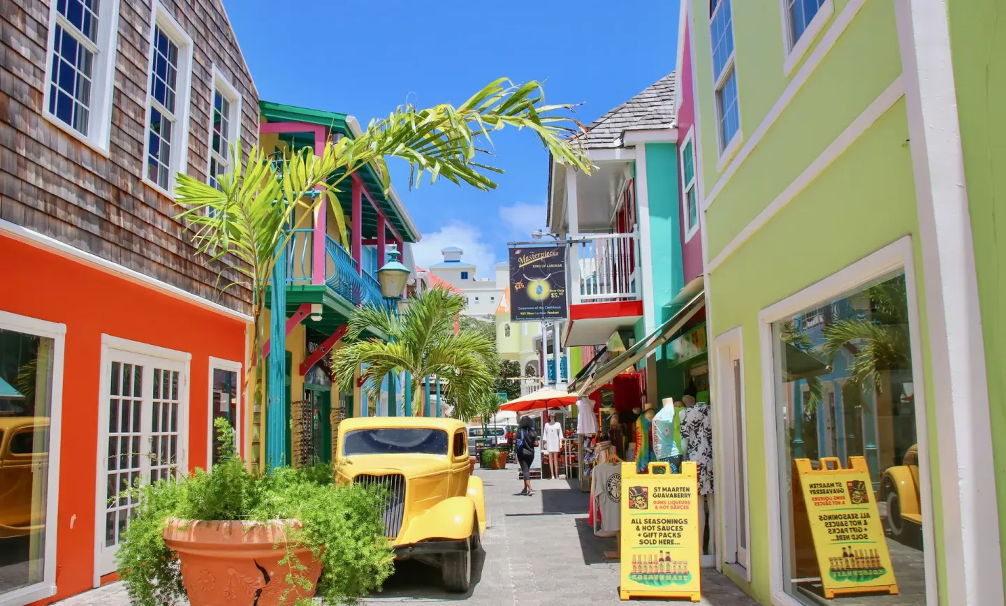 Colorful shopping street near Holland House Beach Hotel with pastel buildings, palms, and a yellow car.