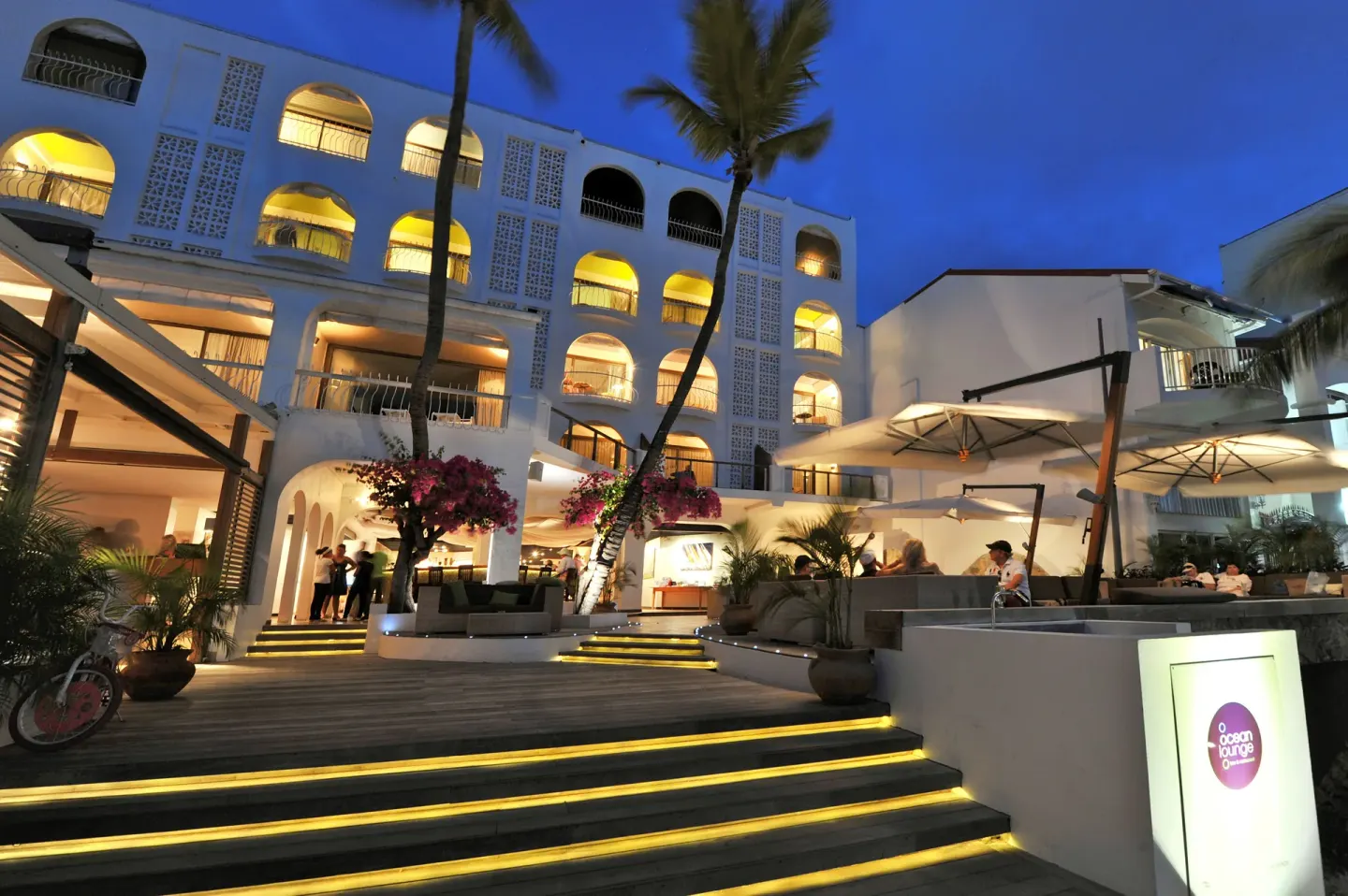 Evening exterior with lit arches, terrace seating, and Ocean Lounge sign at Holland House Beach Hotel.