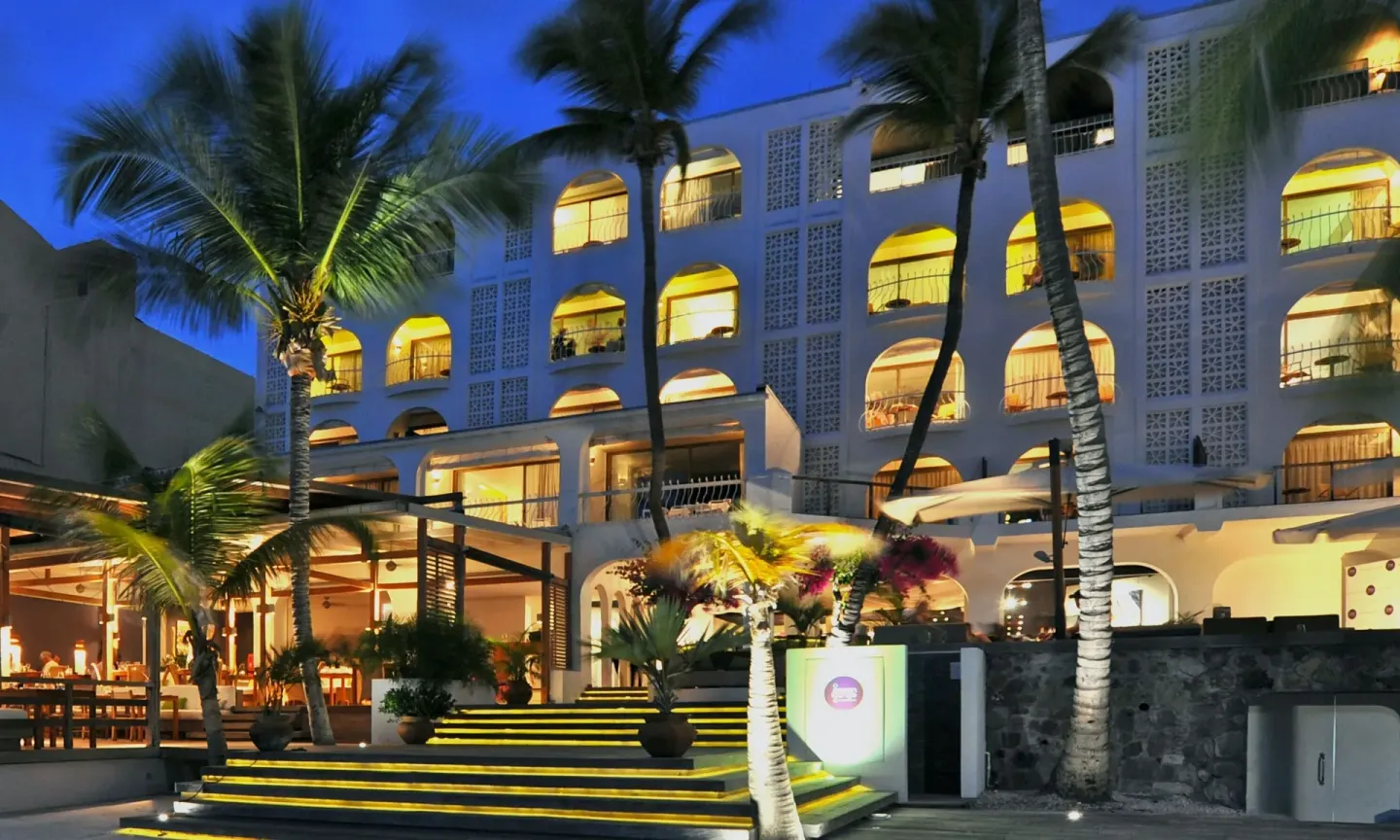 Illuminated exterior of Holland House Beach Hotel with palms, balconies, and terrace steps at night.