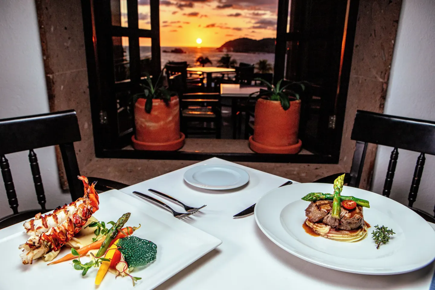 Plated lobster and steak dishes by a sunset window at Hotel Azul Ixtapa Grand Resort Spa.