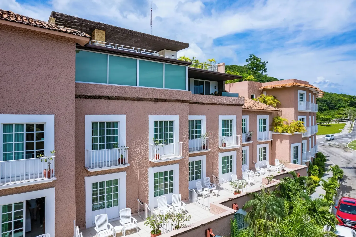 Angled exterior view of Hotel Castillo Huatulco & Beach Club with balconies, terrace chairs, and street.