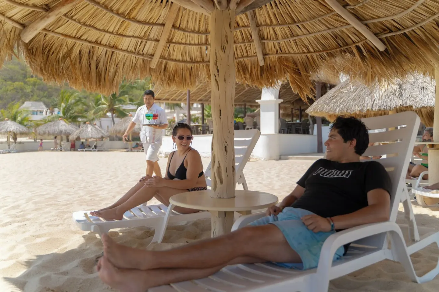 Beach loungers under a thatched umbrella at Hotel Castillo Huatulco & Beach Club.