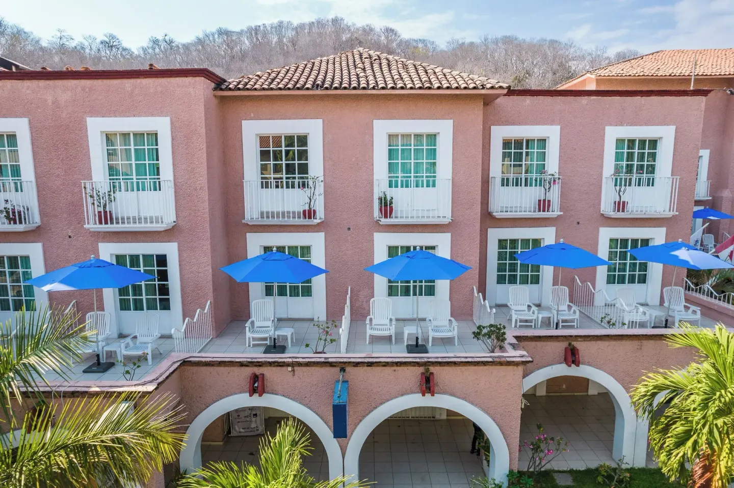 Pink hotel façade with arched walkways and blue patio umbrellas at Hotel Castillo Huatulco & Beach Club.