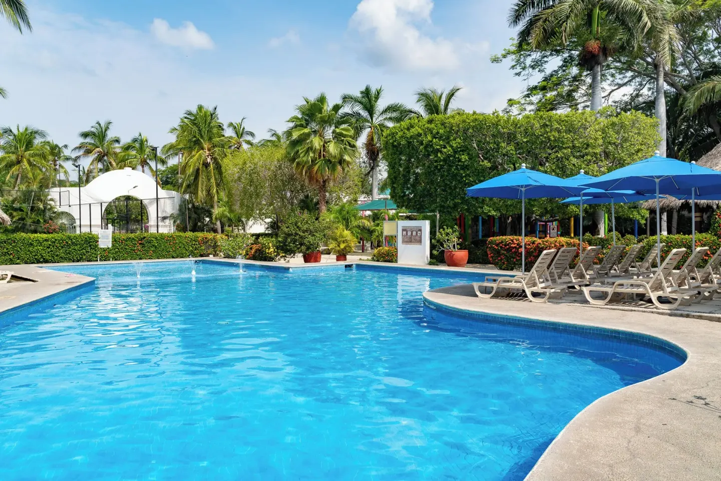 Curved outdoor pool with blue umbrellas and lounge chairs at Hotel Castillo Huatulco & Beach Club.