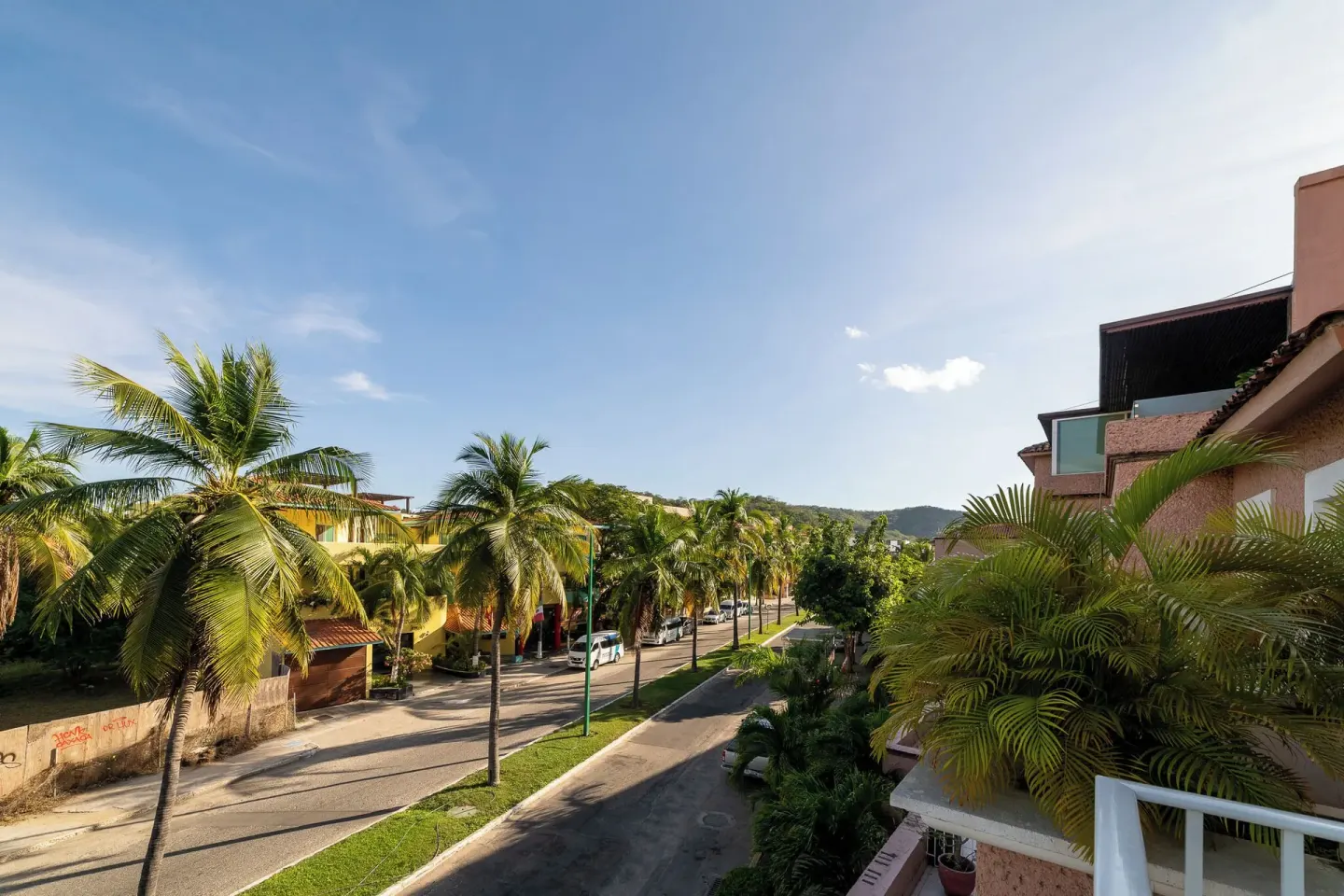 Palm-lined street view from Hotel Castillo Huatulco & Beach Club with nearby buildings and hills.