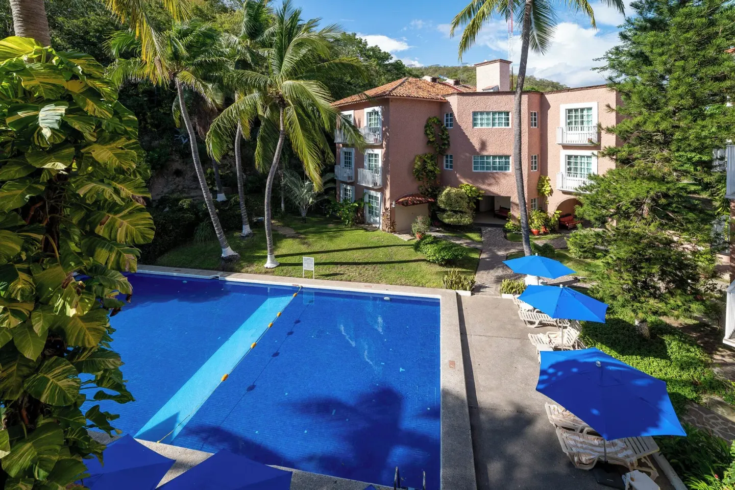 Pool courtyard at Hotel Castillo Huatulco & Beach Club with blue umbrellas, palms, and pink buildings.