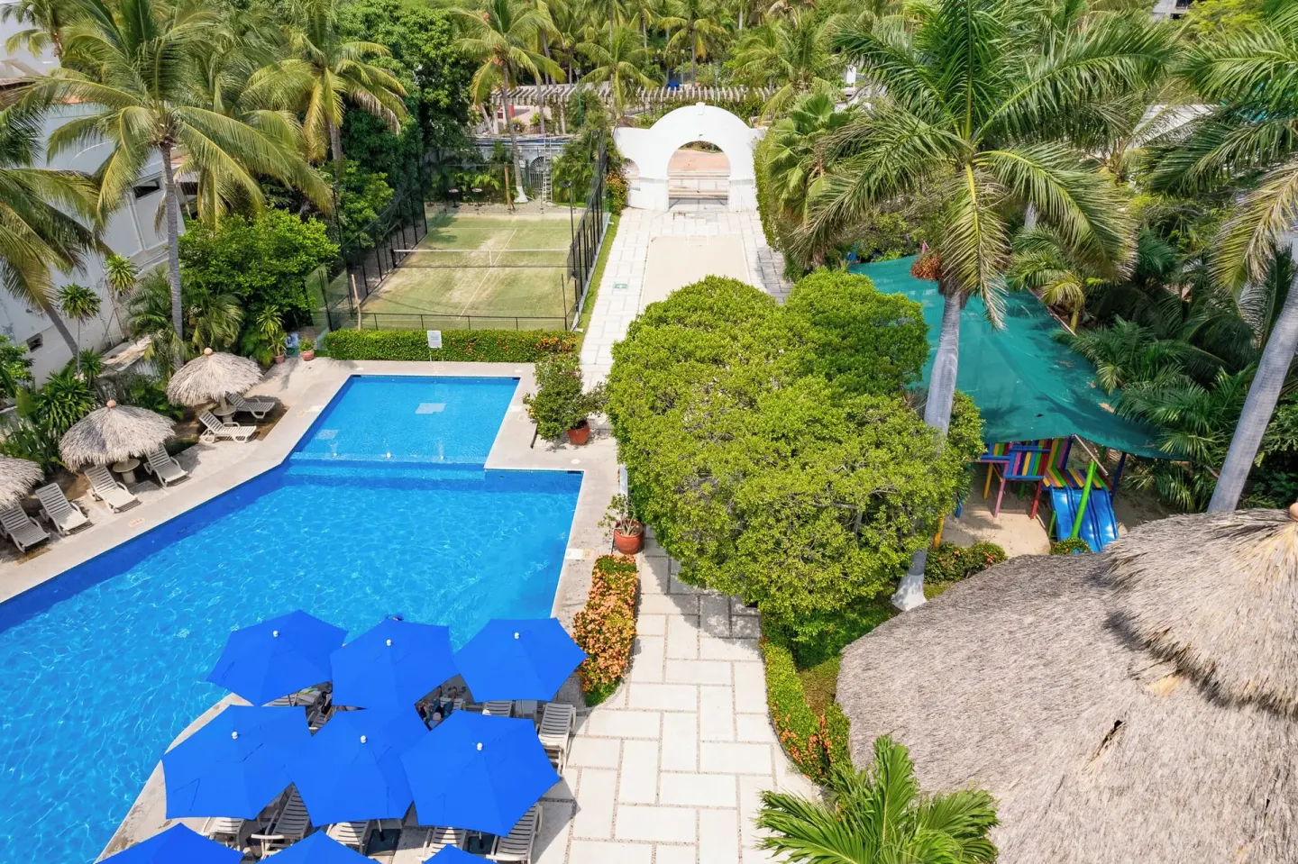 Aerial view of the pool, palms, tennis court, and play area at Hotel Castillo Huatulco & Beach Club.