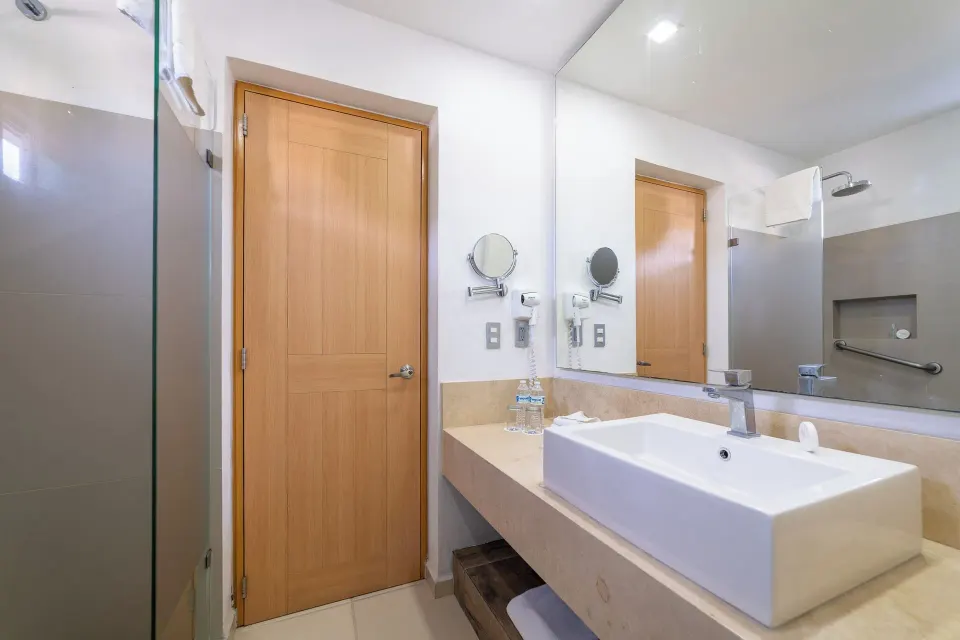 Standard Room bathroom with vanity sink, mirror, glass shower, and wood door at Hotel Castillo Huatulco & Beach Club.