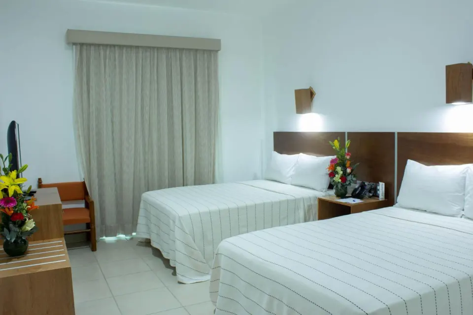 Standard room with two beds, curtains, desk, TV, and flowers at Hotel Castillo Huatulco & Beach Club.