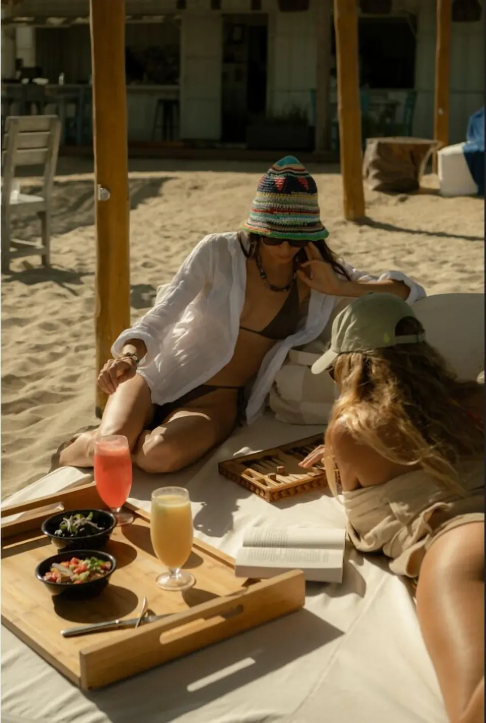 Two guests lounge on a sandy beach daybed with drinks and snacks at Hotel El Ganzo.