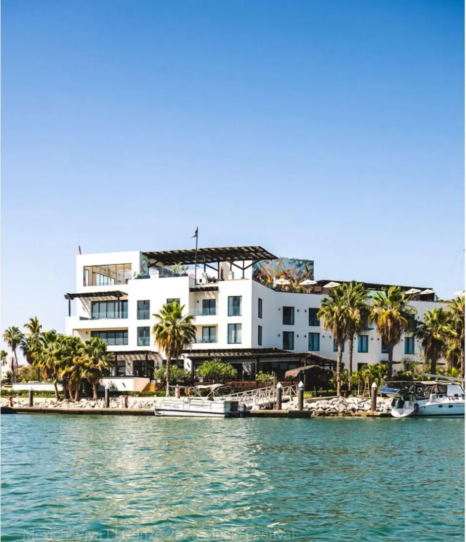 Waterfront exterior of Hotel El Ganzo with palms, marina docks, and boats under a blue sky.