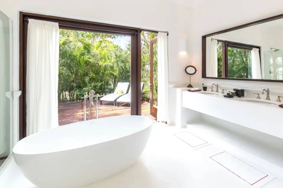 White bathroom with a freestanding bathtub and floor-to-ceiling doors to a leafy deck at Hotel Esencia.