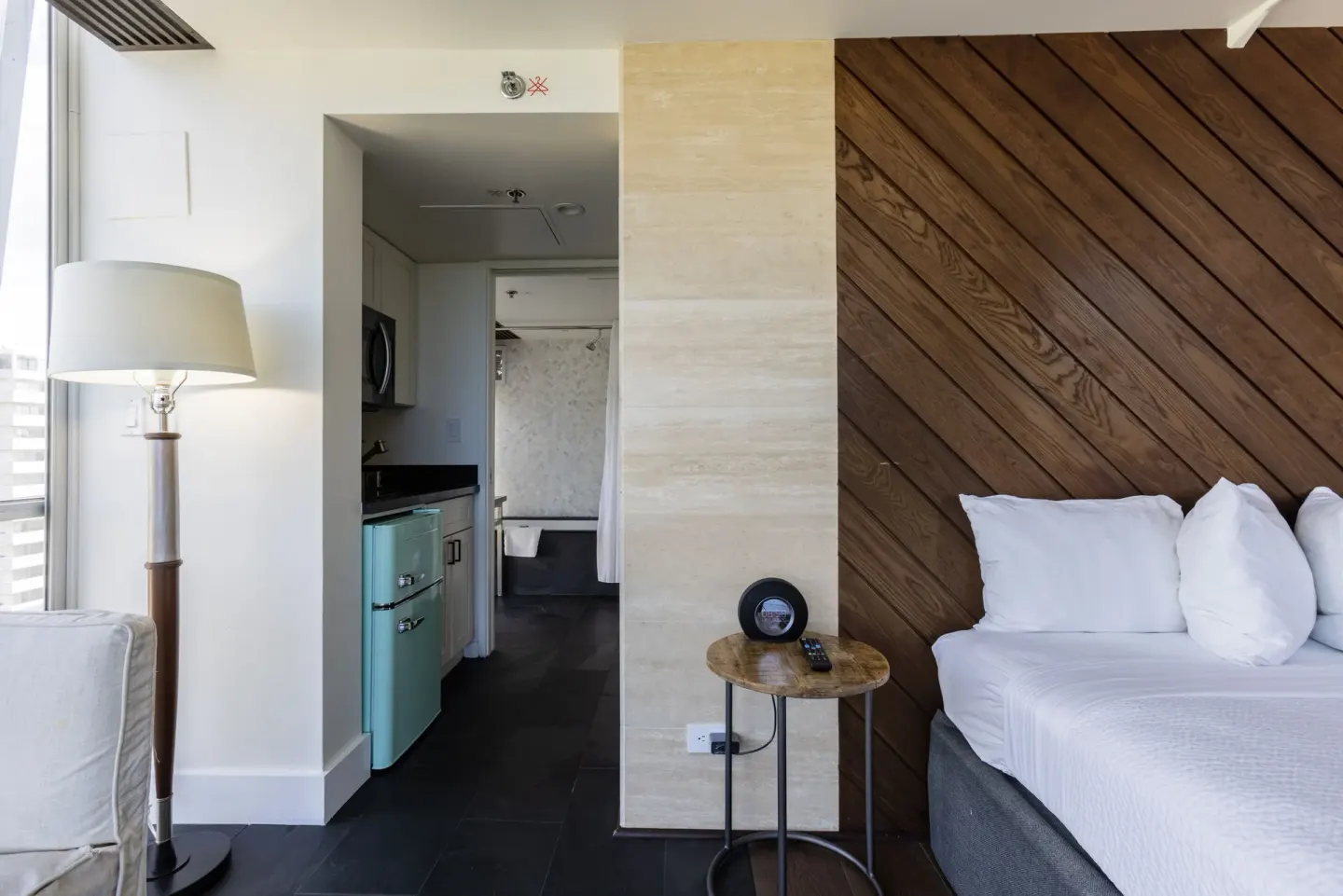 Guest room bed and kitchenette area at Hotel La Croix with wood accent wall and floor lamp.