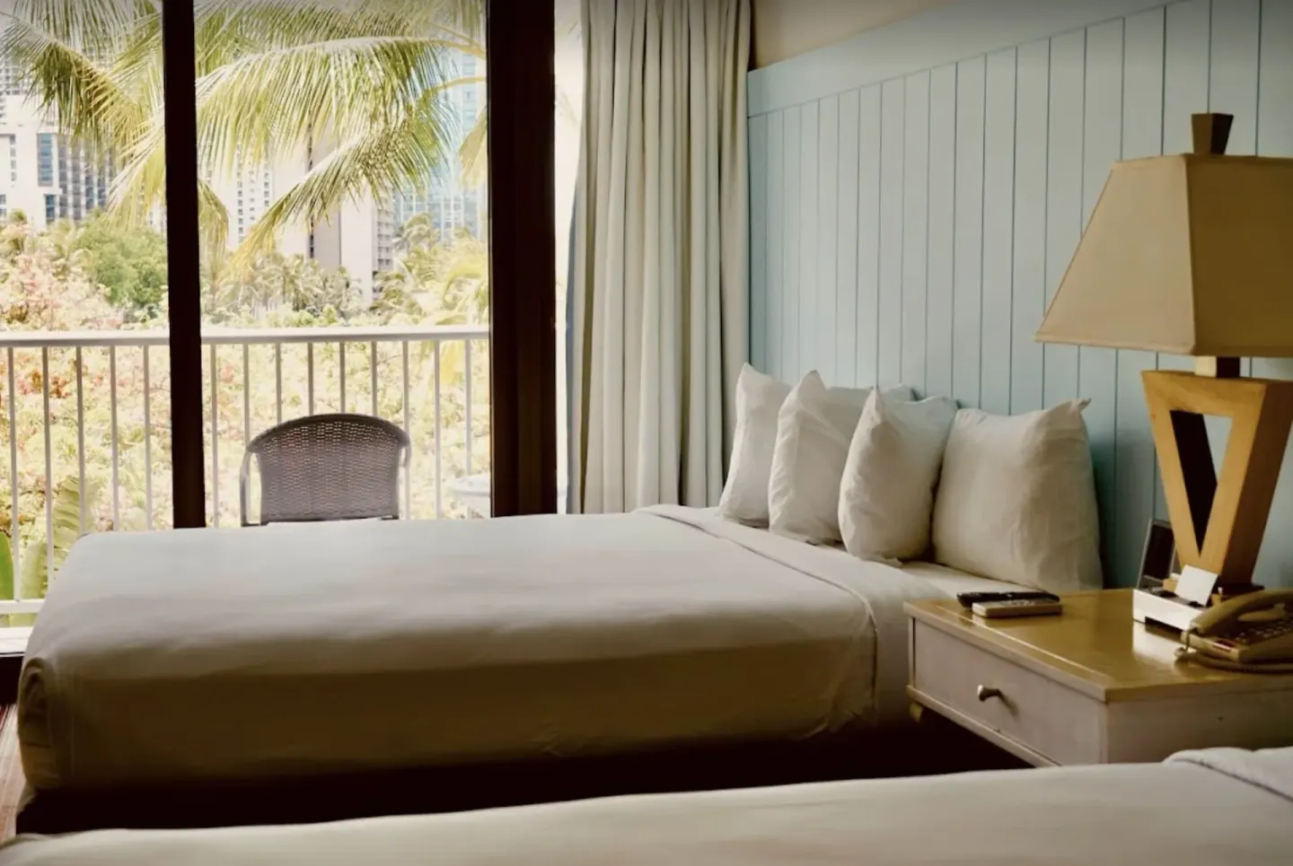 Two-bed guest room at Hotel La Croix with balcony doors, palm view, and bedside lamp.