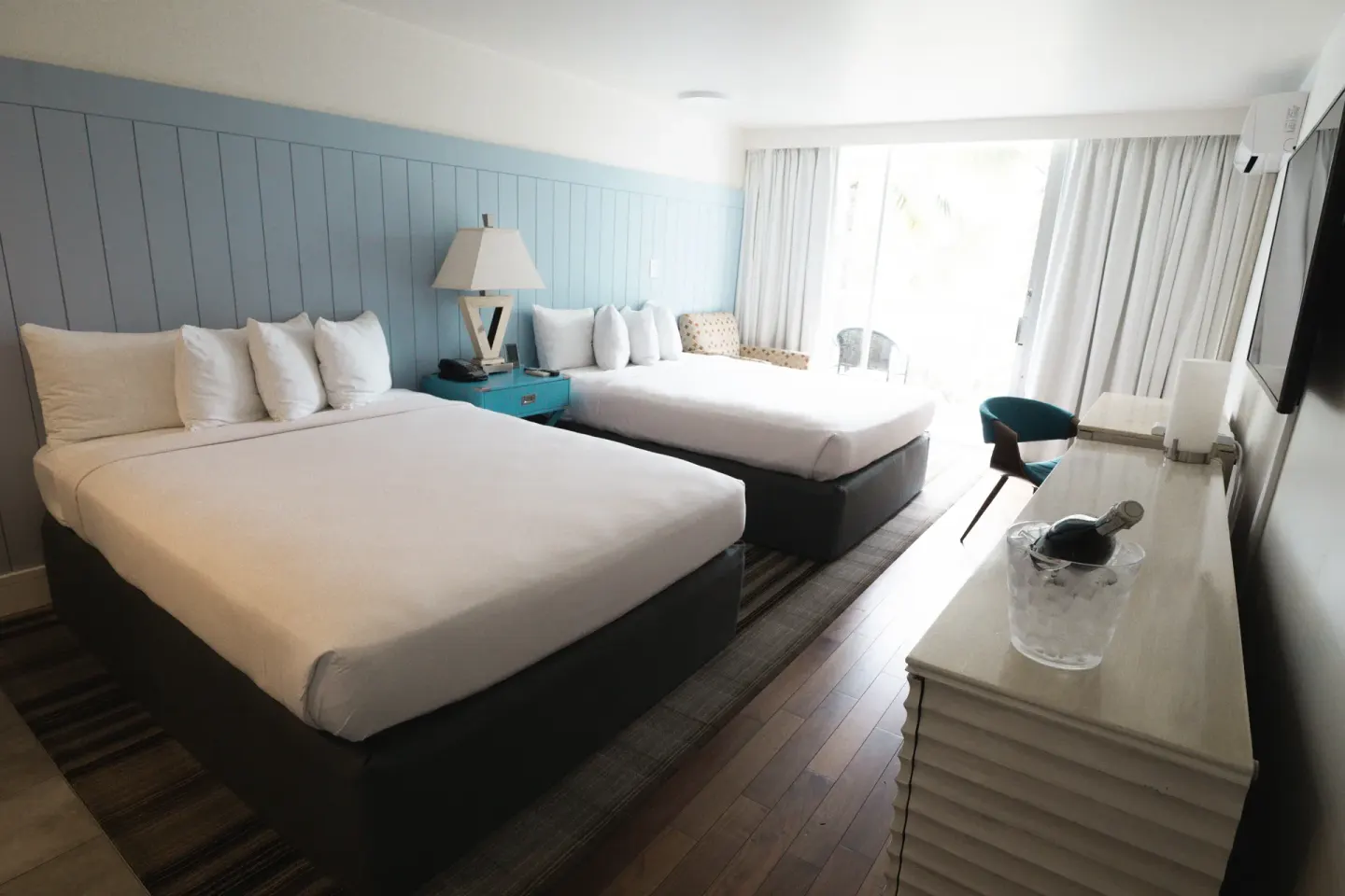 Two-bed guest room at Hotel La Croix with dresser, TV, seating, and bright window.