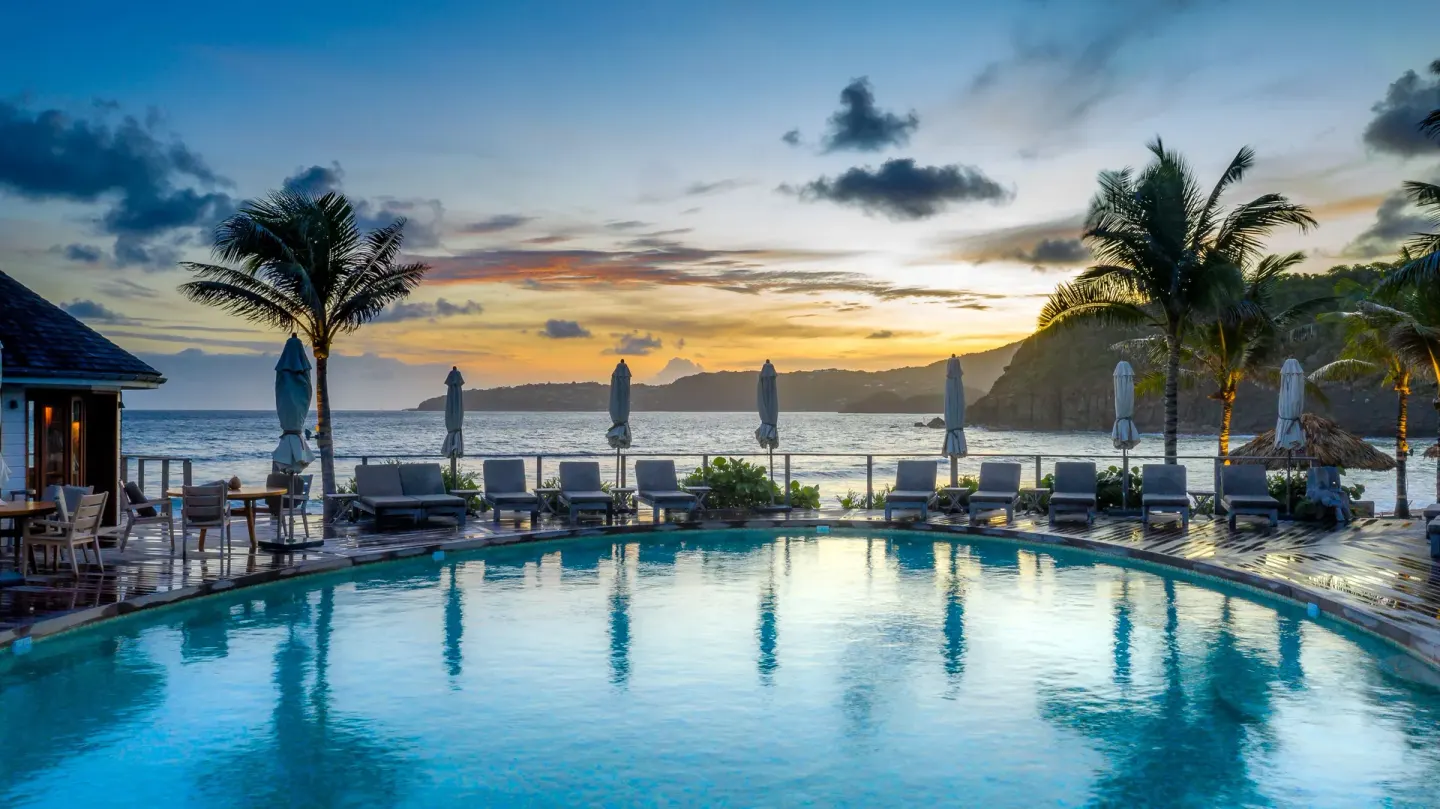 Pool terrace at sunset with loungers, umbrellas, palms, and ocean views at Hotel Manapany.