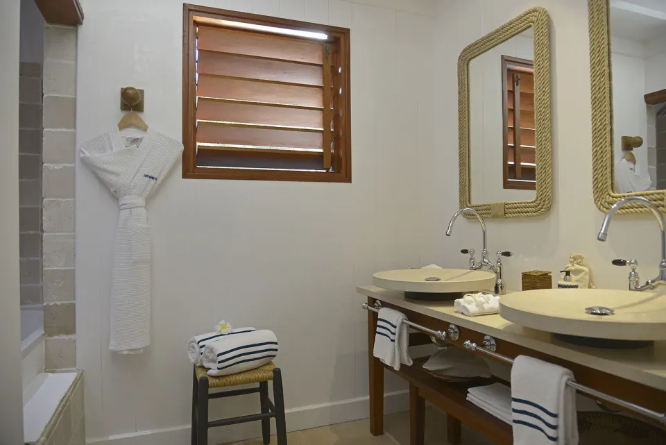 Garden View Room bathroom with twin sinks, rope mirrors, robes, and shutters at Hotel Manapany.